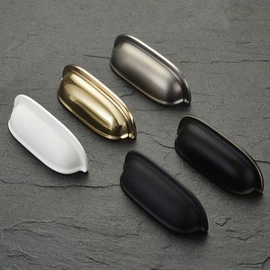 May include: Five cabinet pulls in different finishes: nickel, brass, white, black, and satin nickel. The pulls are all the same shape, with a curved top and bottom.