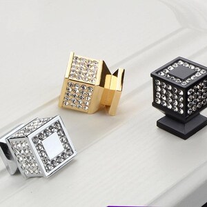 K&ouml;nnte beinhalten: Drei dekorative Schrankkn&ouml;pfe in Gold, Silber und Schwarz. Jeder Knopf hat ein quadratisches Design, das mit klaren Kristallen besetzt ist. Der goldene Knopf ist abgewinkelt, w&auml;hrend die silbernen und schwarzen Kn&ouml;pfe aufrecht stehen.