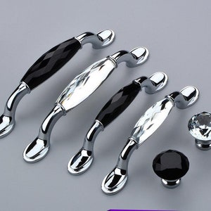 Könnte beinhalten: Vier silberne Schrankknöpfe mit schwarzen und klaren Kristall-Akzenten. Die Knöpfe haben ein geschwungenes, gewölbtes Design.