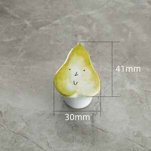 May include: A whimsical pear-shaped ceramic object with a smiling face. The pear is primarily yellow and green, with black details for the eyes and mouth. It measures approximately 1.2 inches wide and 1.6 inches tall. The pear sits atop a small white base.