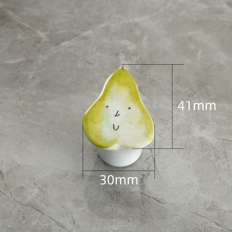 May include: A whimsical pear-shaped ceramic object with a smiling face. The pear is primarily yellow and green, with black details for the eyes and mouth. It measures approximately 1.2 inches wide and 1.6 inches tall. The pear sits atop a small white base.