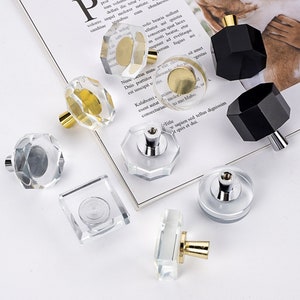 Pode incluir: Conjunto de botões decorativos para armários em várias formas e tamanhos. Os botões são feitos de vidro transparente com detalhes dourados e prateados. Alguns botões são octogonais, enquanto outros são quadrados ou redondos.