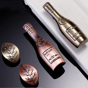 May include: Two antique-style bottle-shaped cabinet door pulls with a leaf-shaped design. The pulls are made of metal and have a distressed finish. The bottle pulls have the words "Remy Martin" embossed on them.