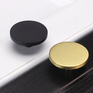 May include: Two round cabinet knobs, one black and one gold. The knobs are made of metal and have a smooth, polished finish.