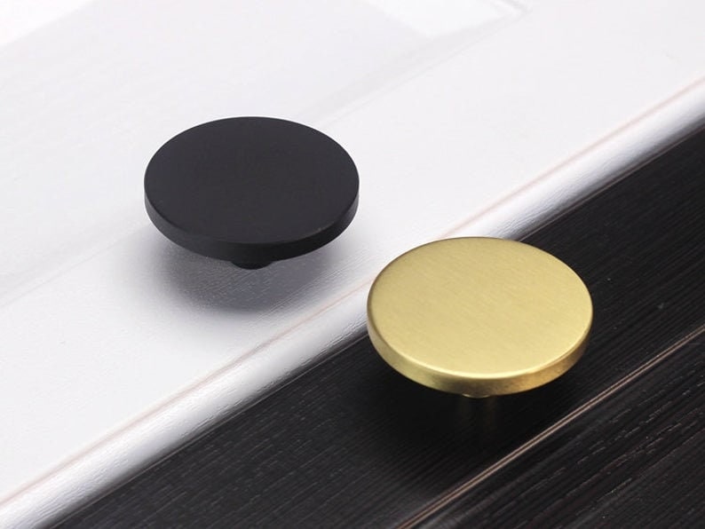 May include: Two round cabinet knobs, one black and one gold. The knobs are made of metal and have a smooth, polished finish.