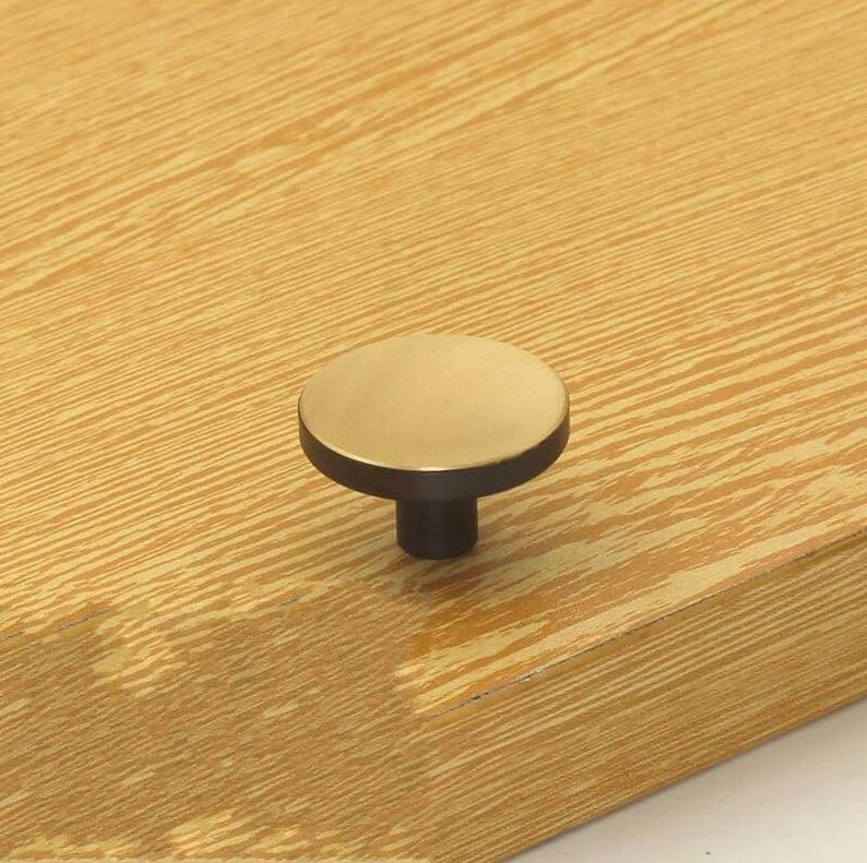 May include: A round, gold-coloured cabinet knob with a black base. The knob is mounted on a light brown wooden surface.