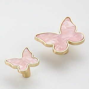 May include: Two gold butterfly-shaped cabinet knobs with pink marbled accents. The larger knob is approximately 5 cm wide and the smaller knob is approximately 2.5 cm wide.