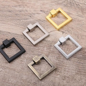 May include: Four square cabinet knobs in different finishes: black, silver, gold, and antique brass. Each knob has a square shape with a small loop handle.