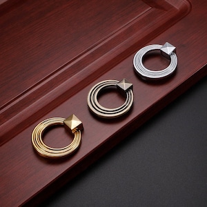 Könnte beinhalten: Drei dekorative Schrankknöpfe mit Ringdesign. Die Knöpfe sind aus Metall und haben eine goldene, silberne und bronzene Oberfläche.