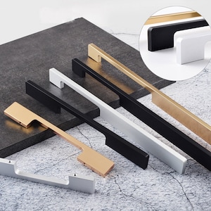 Puede incluir: Cinco tiradores de armario en diferentes acabados: dorado, negro, blanco y plateado. Los tiradores son todos del mismo tamaño y forma, con una base rectangular y una manija curva.