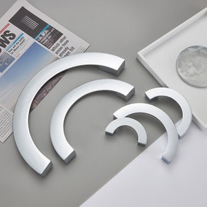 May include: Set of four silver-toned arched cabinet pulls in various sizes. The pulls are arranged in a semi-circle on a gray surface.