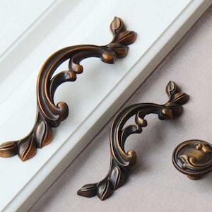 Peut inclure: Ensemble de quincaillerie de meuble de couleur bronze orné. L'ensemble comprend deux poignées incurvées avec des détails en forme de feuilles et un bouton rond. Le matériel est monté sur une surface blanche.