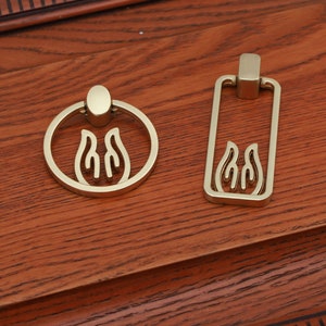 May include: Two gold-toned cabinet pulls with a flame design. One pull is a ring and the other is a rectangle.