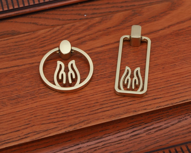 May include: Two gold-toned cabinet pulls with a flame design. One pull is a ring and the other is a rectangle.