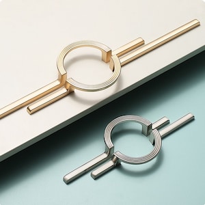 Op de afbeelding: Twee kastgrepen met een modern design. De ene is goudkleurig, de andere zilverkleurig. Elke greep heeft een cirkelvormige handgreep met een gegroefde textuur, verbonden met twee rechte staven. De grepen staan tegen een lichte achtergrond.