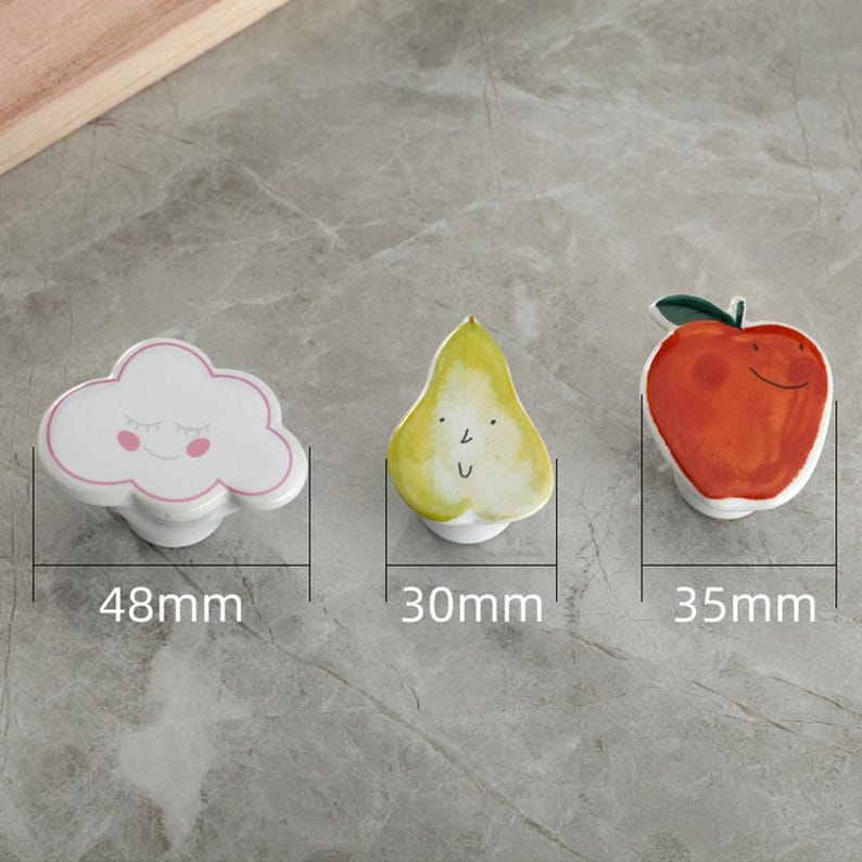 May include: Three ceramic cabinet knobs: a white cloud with pink accents (48mm), a yellow pear with a face (30mm), and a red apple with a green stem (35mm). These decorative knobs are designed for furniture.