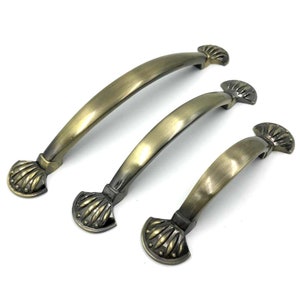 Może przedstawiać: Trzy antyczne mosiężne uchwyty do drzwi szafki z ozdobnymi końcówkami w kształcie muszli. Uchwyty są w różnych rozmiarach.