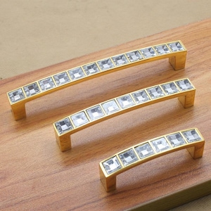 Peut inclure: Trois boutons de porte en or avec une rangée de strass en cristal transparent. Les boutons sont de tailles différentes et sont disposés en rangée sur une surface en bois.