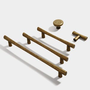 Puede incluir: Conjunto de cuatro tiradores de armario de oro antiguo. Tres son tiradores largos y rectos con una textura similar a la madera. Uno es un pequeño pomo redondo. El cuarto es un pequeño tirador en forma de T.