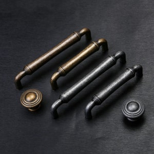 Puede incluir: Cuatro tiradores de armario de estilo antiguo en dos acabados: latón y plata. Los tiradores tienen todos el mismo diseño, con una forma redondeada ligeramente curvada y un anillo decorativo en la parte superior. Dos de los tiradores se muestran con pomos a juego.