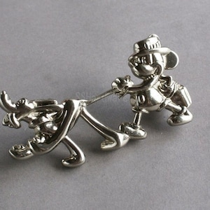 May include: Silver-toned pin featuring a cartoon character pulling another cartoon character on a rope. The character at the front is wearing a hat and has a large nose. The character at the back is holding onto the rope with its mouth.