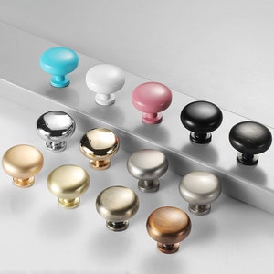 May include: Assortment of 12 cabinet knobs in various finishes, including gold, silver, black, white, pink, and blue. The knobs are round and have a smooth surface.