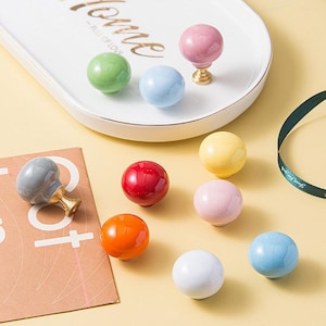 以下が含まれることがあります： 薄い青、ピンク、黄色、赤、オレンジ、グレーなど、さまざまな色をしたセラミック製のキャビネットノブ10個の詰め合わせ。ノブは丸型で、滑らかな仕上げになっています。