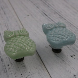 May include: Two ceramic owl drawer pulls, one in light green and one in light blue. Both have a textured surface and black metal mounting hardware.