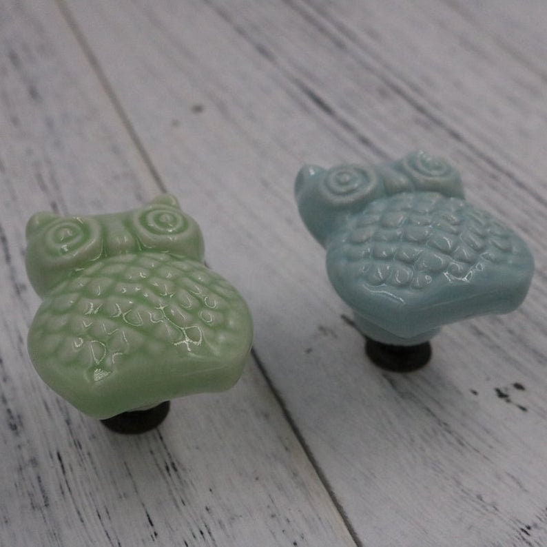 May include: Two ceramic owl drawer pulls, one in light green and one in light blue. Both have a textured surface and black metal mounting hardware.