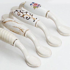 Op de afbeelding: Vier witte keramische kastknoppen met gouden rand. Elke knop heeft een ander decoratief ontwerp, waaronder bloemmotieven, marmer en draaiende patronen.