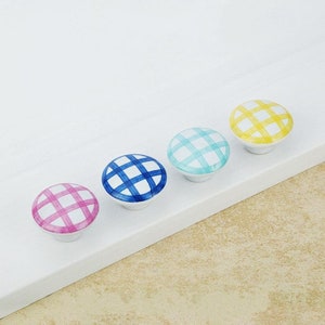 May include: Four ceramic cabinet knobs with a white base and colourful plaid patterns in pink, blue, turquoise, and yellow. The knobs are round and have a glossy finish, ideal for adding a pop of colour to furniture.