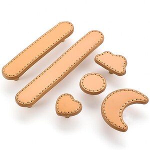 May include: Set of six leather cabinet pulls in various shapes, including a heart, a crescent moon, a cloud, and two rectangular pulls. The pulls are all a light brown colour and have a stitched edge.