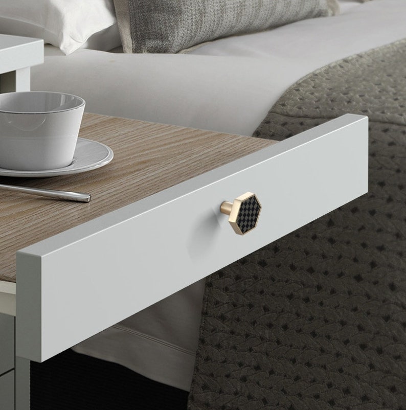 Pu&ograve; includere: Primo piano di un cassetto con una maniglia esagonale nera e dorata. Il cassetto &egrave; in legno bianco ed &egrave; aperto, rivelando una superficie in legno marrone sottostante.