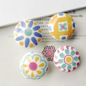 Pode incluir: Quatro puxadores redondos decorativos para armários com desenhos florais coloridos. Os puxadores apresentam padrões em tons de azul, amarelo, rosa e verde-azulado. Os puxadores são feitos de cerâmica ou material semelhante.