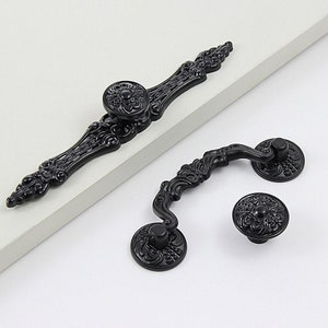 Könnte beinhalten: Zwei schwarze, verzierte Metall-Schrankgriffe. Einer ist ein langer Griff mit einem runden Knopf in der Mitte. Der andere ist ein kürzerer Griff mit zwei runden Knöpfen.