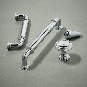 May include: Four polished chrome cabinet hardware pieces: two pulls, one knob, and one small decorative piece. The pulls are both curved and have a cylindrical design. The knob is round with a raised edge. The decorative piece is a small, rounded knob.