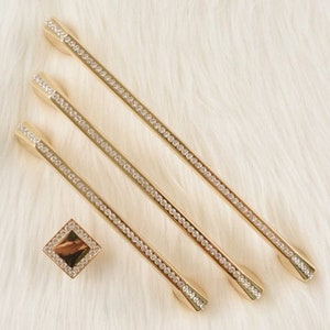 May include: Three gold cabinet pulls with a row of clear rhinestones. The pulls are on a white fuzzy surface.