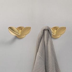 Può includere: Due ganci dorati a forma di foglia montati su una parete. Un gancio ha un asciugamano grigio appeso.