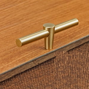 Pode incluir: Uma puxador de armário em forma de T de cor dourada com bordas arredondadas.