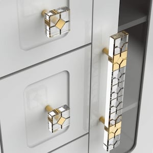Op de afbeelding: Close-up van witte kastdeuren met decoratieve hardware. De kast heeft vierkante knoppen en een lange handgreep, allemaal met een helder, geometrisch ontwerp en gouden accenten. De hardware voegt een vleugje elegantie toe aan de moderne kasten.