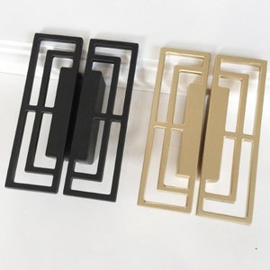 May include: Two cabinet pulls with a geometric design. One pull is black and the other is gold.