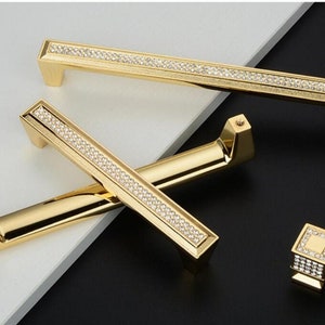 May include: Three gold cabinet knobs with a diamond-like pattern. The knobs are in various sizes and shapes. The largest knob is rectangular and has a long, thin handle. The smallest knob is square and has a flat top. The middle knob is a combination of the two, with a rectangular base and a square top.