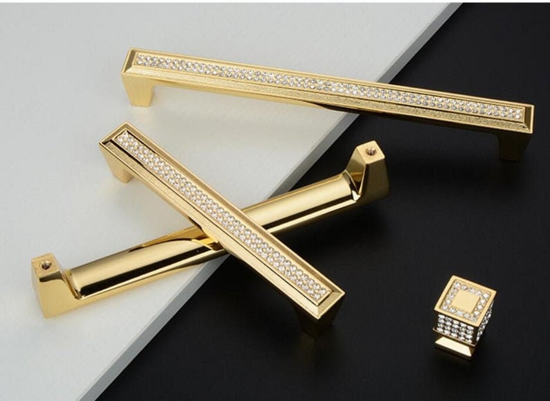 May include: Three gold cabinet knobs with a diamond-like pattern. The knobs are in various sizes and shapes. The largest knob is rectangular and has a long, thin handle. The smallest knob is square and has a flat top. The middle knob is a combination of the two, with a rectangular base and a square top.