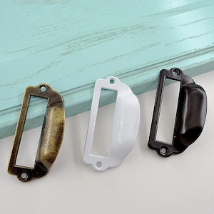 Puede incluir: Tres tiradores de cajón en diferentes acabados: dorado, blanco y negro. Cada tirador tiene un portaetiquetas rectangular con bordes redondeados.