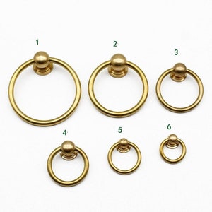 May include: A collection of six brass ring pulls in varying sizes. Each pull features a circular ring attached to a round base. The pulls are arranged on a white surface, showcasing their different dimensions and designs.