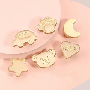 May include: Six gold-toned metal drawer pulls with different designs: a car, a paw print, a crescent moon, a heart with the word "LOVE" inscribed, a star, and a bear.