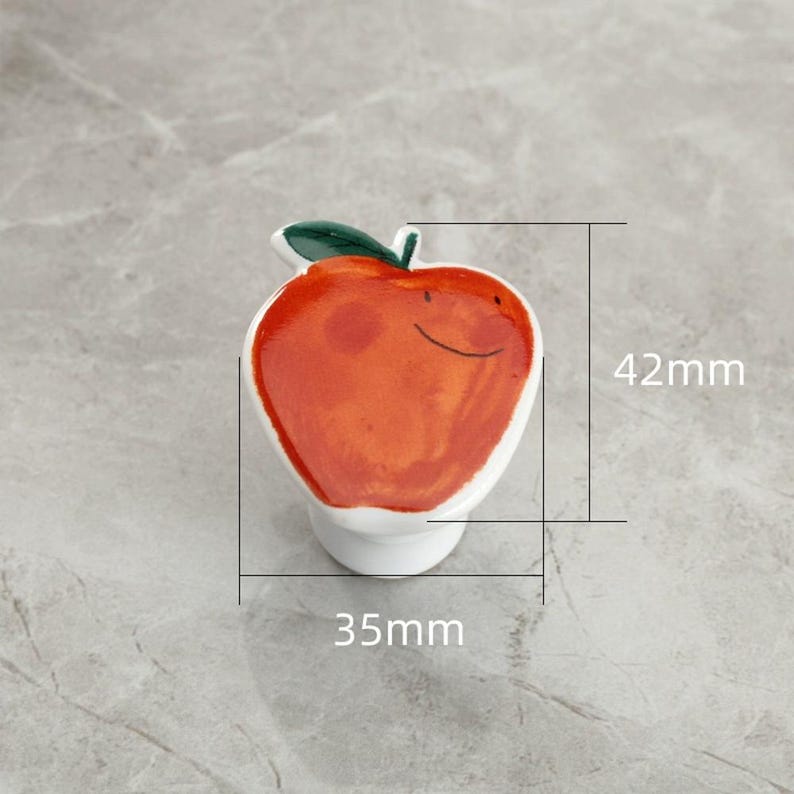 May include: A ceramic apple-shaped cabinet knob. The knob is painted with a red apple design, a green leaf, and a white base. The knob measures 1.4 inches in height and 1.4 inches in width.