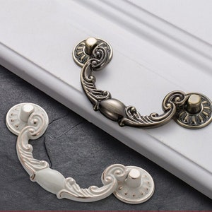 以下が含まれることがあります： 装飾的な曲線デザインの装飾的なキャビネットプル2つ。1つのプルはアンティークブラスで、中心が白。もう1つは白で、中心が白です。