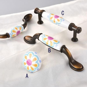 May include: Three ceramic cabinet knobs with a floral design in shades of yellow, blue, pink, and white. The knobs have a bronze finish.
