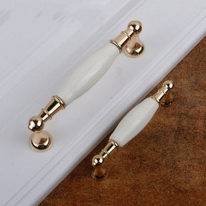 May include: Two white ceramic cabinet door pulls with gold metal accents. The pulls are different sizes.